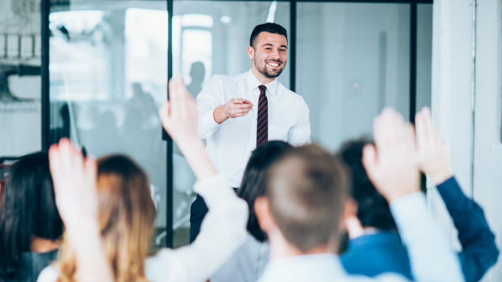 A smiling man in a shirt and tie leads a workshop, engaging with attentive participants who are raising their hands to answer a question.